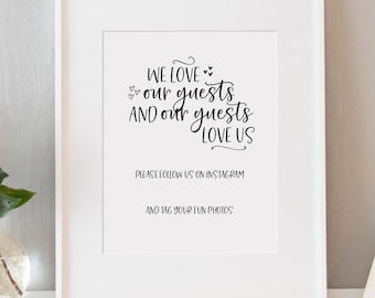 May include: A white framed print with black text that reads "WE LOVE our guests AND our guests LOVE US" and "PLEASE FOLLOW US ON INSTAGRAM AND TAG YOUR FUN PHOTOS". The print is on a white shelf with a pink flower in a clear vase, a white letter A, and a small potted plant.
