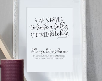 May include: A white framed print with black text that reads "WE STRIVE to have a fully STOCKED kitchen Please let us know IF YOU RUN OUT OF SOMETHING OR IF SOMETHING IS MISSING". The print is sitting on a gray surface with a wooden triangle shelf and a purple mug.