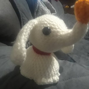 May include: A handmade white crocheted dog with a long snout, black eyes, and a red collar. The dog is holding an orange ball in its mouth. The dog is a character from the movie "The Nightmare Before Christmas".