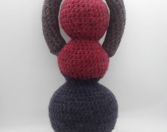 May include: A handmade, abstract crochet sculpture in shades of burgundy, dark gray, and brown. The piece features a dark gray, teardrop-shaped base, topped by two burgundy spheres, and a brown, circular halo. The texture is chunky and tactile.