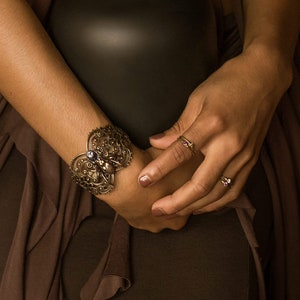 Puede incluir: Una persona con un vestido marrón sostiene un jarrón negro con una planta de helecho dentro. La persona lleva un brazalete dorado con un diseño decorativo y dos anillos en los dedos.