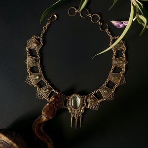 May include: A gold-toned necklace with a large, faceted, clear gemstone pendant. The necklace is made of intricate metalwork and has a delicate chain. The necklace is lying on a black surface with a snake skin pattern.