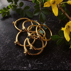 以下が含まれることがあります： 緑と紫の宝石が付いた金色のリング5つのセット。リングは互いに重ねられ、円形に配置されています。