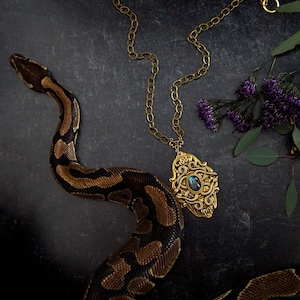 May include: A gold-toned pendant necklace with an ornate design and a blue-green stone is displayed. A brown and black snake is coiled around the necklace. Purple flowers and greenery are in the background. The necklace has a chain.