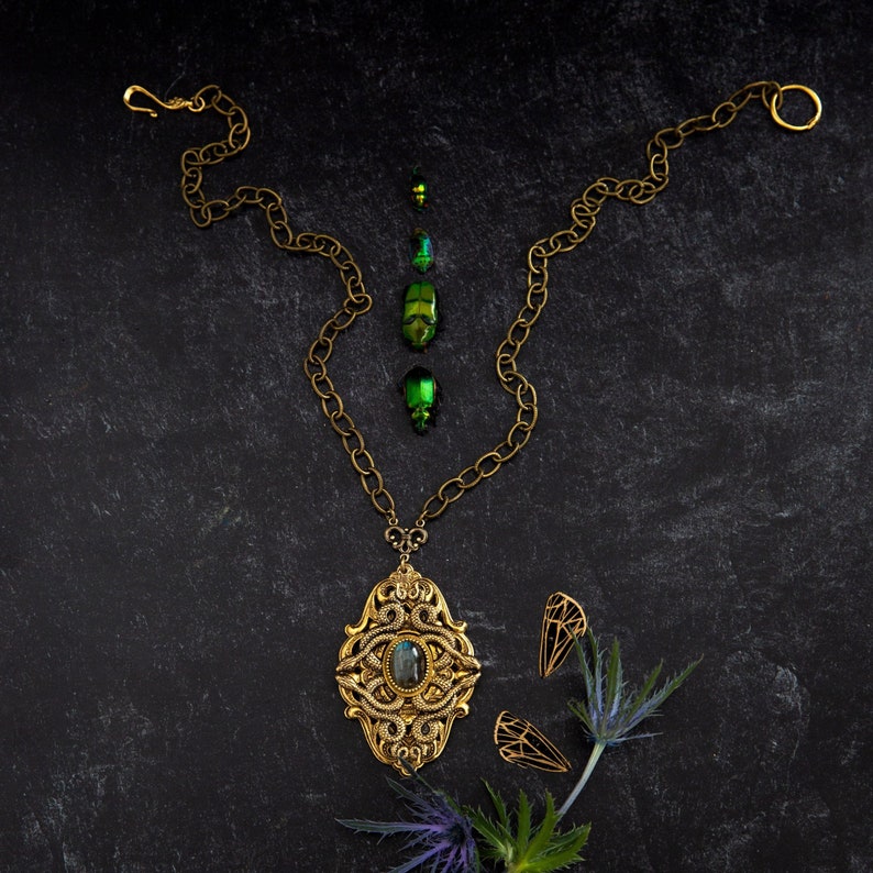 May include: An antique gold-toned necklace with an ornate pendant featuring a central stone. The necklace is accompanied by matching earrings and iridescent green beetle shells, arranged on a dark textured surface. The chain is a chunky link style.
