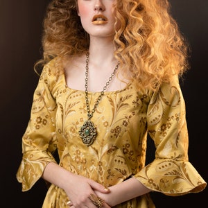 May include: A woman wearing a gold-colored dress with a floral pattern and a square neckline. She is wearing a long, ornate necklace with a pendant and a large ring. Her hair is styled in voluminous curls, and she has gold-colored makeup.