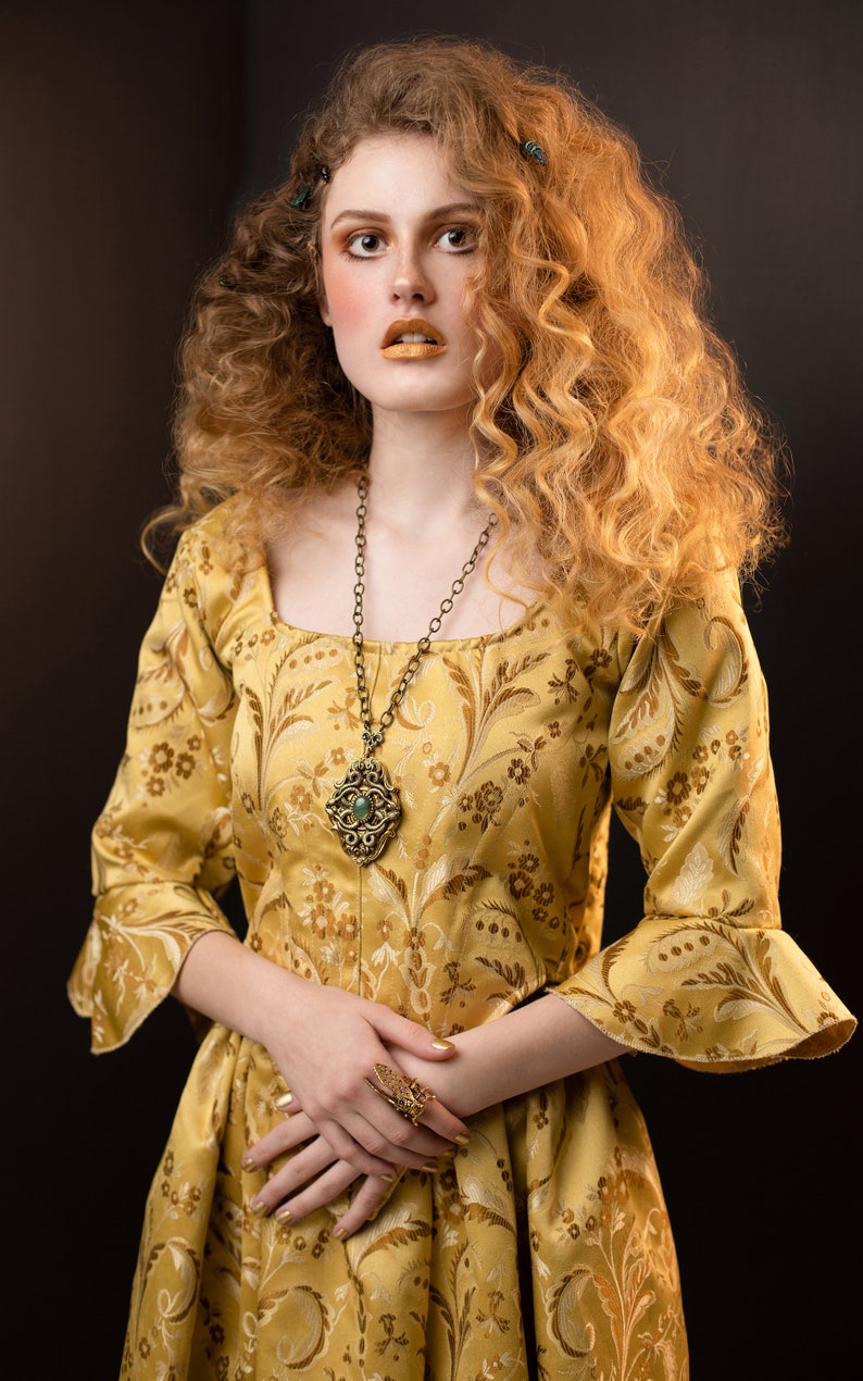 May include: A woman wearing a gold-colored dress with a floral pattern and a square neckline. She is wearing a long, ornate necklace with a pendant and a large ring. Her hair is styled in voluminous curls, and she has gold-colored makeup.