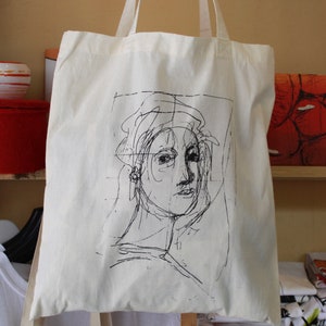 Puede incluir: Una bolsa de tela blanca con un dibujo de línea negra de una cara en la parte delantera. El dibujo es de estilo simple y esquemático, y la persona lleva un sombrero.