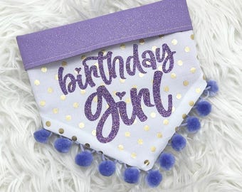 Könnte beinhalten: Ein weißes Hunde-Bandana mit goldenen Tupfen und dem Schriftzug "birthday girl" in glitzerndem Lila. Der obere Rand ist aus lila Glitzerstoff und die Unterseite hat blaue Pompons.