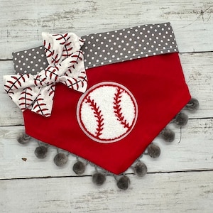 May include: Red dog bandana with a baseball applique and a white and red baseball-themed bow. The bandana has a gray polka dot trim and gray pom-pom trim along the bottom edge. The baseball applique is white with red stitching.