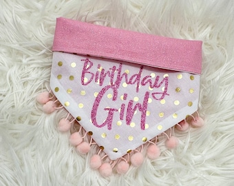 May include: A pink and white dog bandana with the text "Birthday Girl" in glittery pink script. The bandana has gold polka dots and is trimmed with pink pom-poms. The top band is a glittery pink fabric.