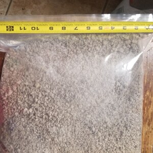 May include: A clear plastic bag filled with light-colored granular material. A yellow measuring tape is placed across the top of the bag, showing measurements in inches. The bag appears to be sealed.