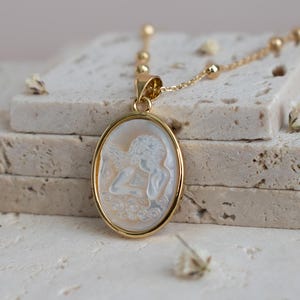 Puede incluir: Un collar con colgante ovalado en tono dorado que presenta un camafeo blanco tallado con una figura de perfil. El colgante cuelga de una delicada cadena dorada con peque&ntilde;as cuentas esf&eacute;ricas espaciadas a lo largo de la cadena de eslabones.