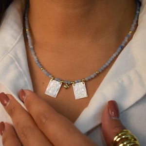 May include: A delicate necklace featuring small, light blue beads and gold accents. Two rectangular pendants with a white, textured design hang from the necklace. The necklace is worn over a white collared shirt.