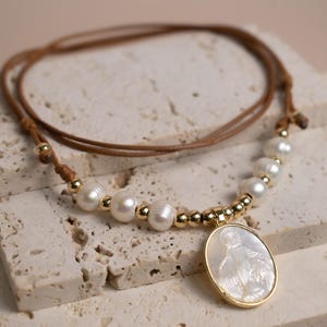 K&ouml;nnte beinhalten: Eine Halskette mit braunem Band, wei&szlig;en S&uuml;&szlig;wasserperlen und kleinen goldfarbenen Perlen. Das Herzst&uuml;ck ist ein ovaler Anh&auml;nger mit einer Einlage aus wei&szlig;em Perlmutt in einem goldfarbenen Rahmen, der eine Figur im Relief darstellt.