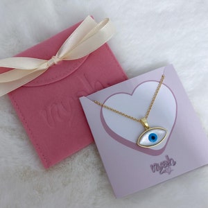 May include: A gold-tone chain necklace with an eye-shaped pendant featuring a white mother-of-pearl inlay and a blue center pupil. The necklace is displayed on a light purple card with a heart graphic and the text nyah, next to a pink pouch.
