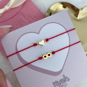 May include: Two red string bracelets with gold charms. One charm is a heart and the other is an infinity symbol. The bracelets are displayed on a white card with the word "nyah" written in cursive.