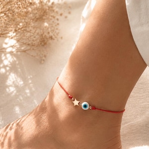 Peut inclure: Une chevillère en cordon rouge ornée d'une petite breloque étoile dorée et d'une perle ronde blanche en forme d'œil porte-bonheur avec un centre bleu et noir. Le cordon est agrémenté de petites perles dorées de chaque côté des breloques.