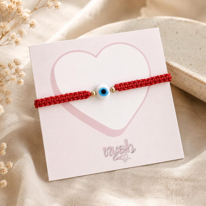 K&ouml;nnte beinhalten: Ein rotes, geflochtenes Armband mit einer zentralen, wei&szlig;-blau-schwarzen Perle in Form eines b&ouml;sen Blicks und kleinen goldfarbenen Metallakzenten. Das Armband ist auf einer quadratischen wei&szlig;en Karte mit rosa Herzumriss und dem Text nyah befestigt.