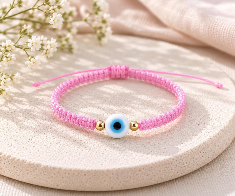 K&ouml;nnte beinhalten: Ein Armband aus rosa geflochtener Kordel mit einer zentralen, runden Glasperle in Form eines Schutzauges in Wei&szlig;, Hellblau und Schwarz, flankiert von zwei kleinen, goldfarbenen Metallperlen. Das Armband hat einen verstellbaren Schiebeknoten.