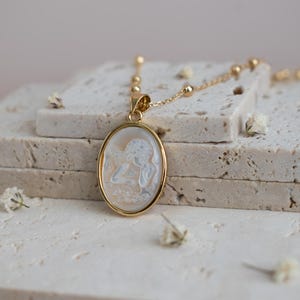 Puede incluir: Un collar con colgante ovalado en tono dorado que presenta un camafeo blanco tallado con una figura de perfil. El colgante cuelga de una delicada cadena dorada con peque&ntilde;as cuentas esf&eacute;ricas espaciadas a lo largo de la cadena de eslabones.