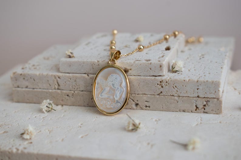 Puede incluir: Un collar con colgante ovalado en tono dorado que presenta un camafeo blanco tallado con una figura de perfil. El colgante cuelga de una delicada cadena dorada con peque&ntilde;as cuentas esf&eacute;ricas espaciadas a lo largo de la cadena de eslabones.