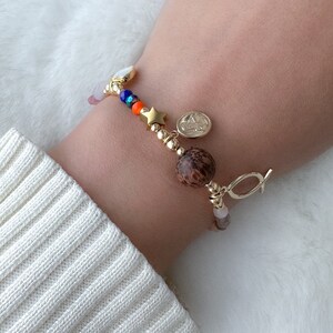 May include: A gold bracelet with a variety of charms, including a star, a wooden bead, and a small religious medal. The bracelet also features pink, blue, and orange beads.