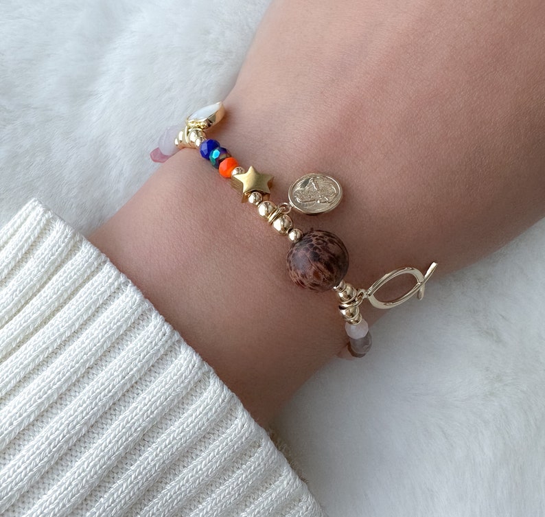 May include: A gold bracelet with a variety of charms, including a star, a wooden bead, and a small religious medal. The bracelet also features pink, blue, and orange beads.