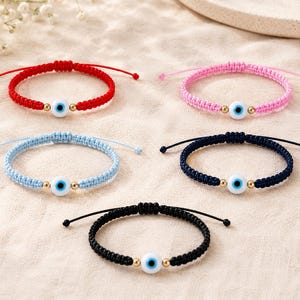 K&ouml;nnte beinhalten: F&uuml;nf verstellbare Armb&auml;nder aus Kordel in Rot, Pink, Hellblau, Dunkelblau und Schwarz. Jedes Armband hat eine zentrale wei&szlig;e Perle mit einem blauen Auge, flankiert von zwei kleinen goldfarbenen Metallperlen auf einem geflochtenen Nylonband.