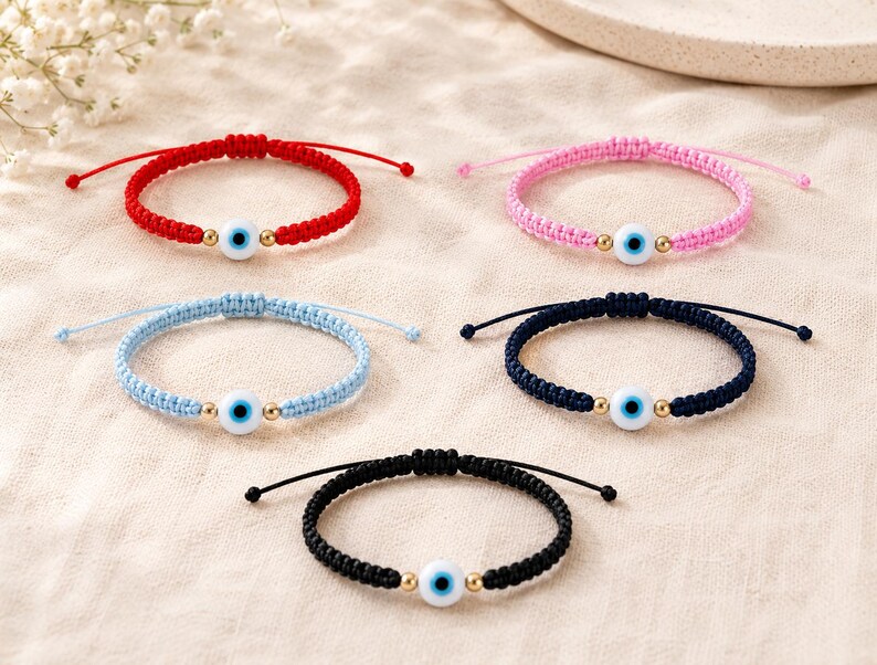 K&ouml;nnte beinhalten: F&uuml;nf verstellbare Armb&auml;nder aus Kordel in Rot, Pink, Hellblau, Dunkelblau und Schwarz. Jedes Armband hat eine zentrale wei&szlig;e Perle mit einem blauen Auge, flankiert von zwei kleinen goldfarbenen Metallperlen auf einem geflochtenen Nylonband.