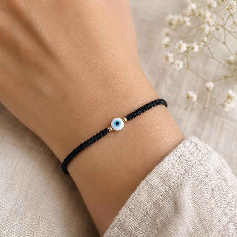 K&ouml;nnte beinhalten: Ein schwarzes Armband aus Kordel mit einer zentralen, runden Schutzaugen-Perle in Wei&szlig;, Blau und Schwarz, die von zwei kleinen goldfarbenen Metallperlen flankiert wird. Das Armband ist geflochten und verf&uuml;gt &uuml;ber einen Schiebeknoten-Verschluss.