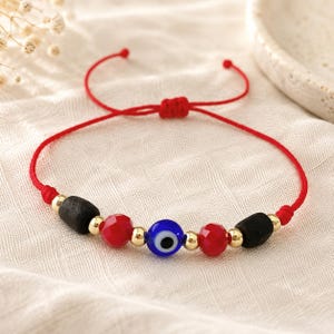 May include: A red cord bracelet featuring a central blue, white, and black evil eye bead, flanked by two red faceted beads, two matte black cylindrical beads, and several small gold-toned metallic spacer beads with an adjustable sliding knot.
