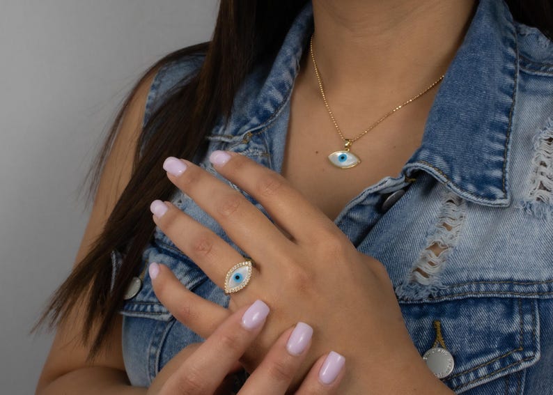 May include: A gold-toned necklace and ring set featuring matching evil eye pendants. Each piece displays a central blue and white eye motif surrounded by a border of small, clear stones. The necklace hangs on a delicate gold chain, while the ring sits on a gold band, both showcasing a polished finish and intricate detailing.