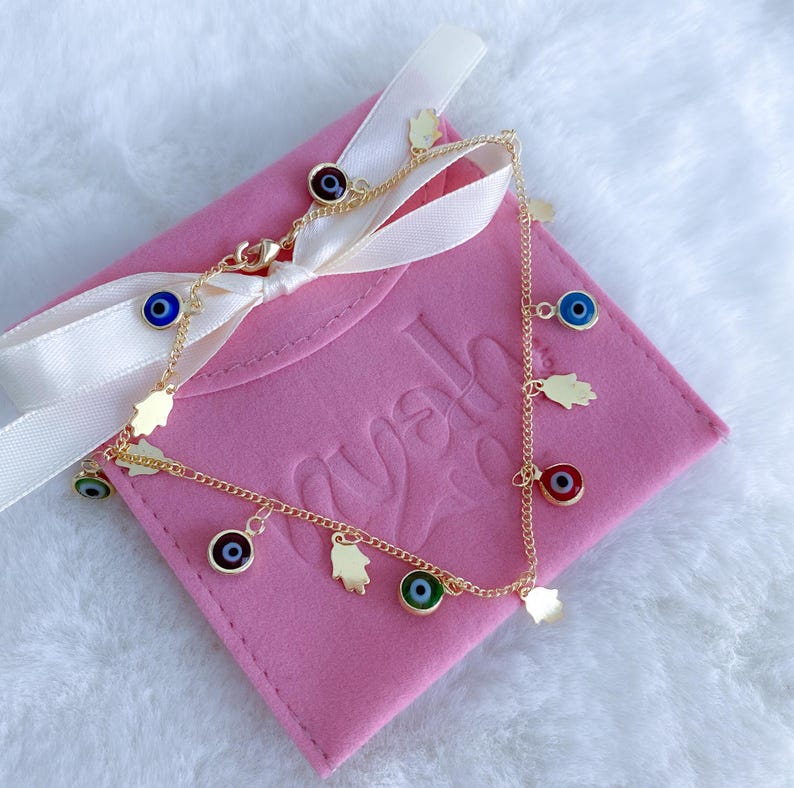 Puede incluir: Tobillera de cadena dorada con m&uacute;ltiples encantos, incluyendo ojos turcos azules, verdes y rojos y encantos de mano de hamsa dorados. La tobillera se muestra en una caja de terciopelo rosa con la palabra "ayah" grabada en la caja.