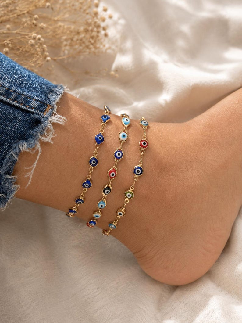 May include: Three gold-tone chain anklets featuring small, circular evil eye charms. The charms are decorated with enamel in various colors, including dark blue, light blue, red, white, and green, arranged in concentric circles on each link.