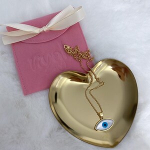 May include: A gold-tone chain necklace with an evil eye pendant featuring a white oval base and a central blue pupil. The necklace rests on a gold-tone heart-shaped dish next to a pink fabric pouch tied with a cream-colored ribbon.