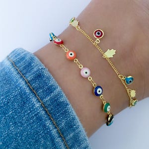 Puede incluir: Dos tobilleras doradas con abalorios de ojo turco de colores. Las tobilleras est&aacute;n superpuestas en el tobillo de una persona. Las tobilleras tienen una cadena delicada y est&aacute;n adornadas con peque&ntilde;os abalorios de mano de hamsa dorados.