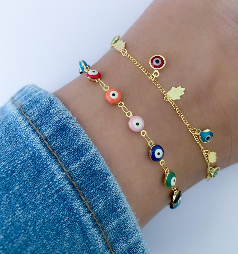 Puede incluir: Dos tobilleras doradas con abalorios de ojo turco de colores. Las tobilleras est&aacute;n superpuestas en el tobillo de una persona. Las tobilleras tienen una cadena delicada y est&aacute;n adornadas con peque&ntilde;os abalorios de mano de hamsa dorados.