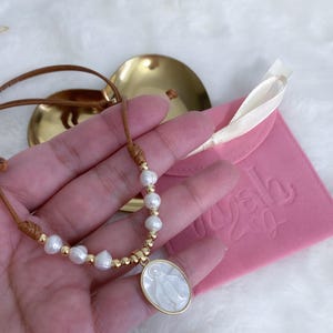 K&ouml;nnte beinhalten: Eine Halskette mit brauner Kordel, kleinen wei&szlig;en S&uuml;&szlig;wasserperlen und goldfarbenen Perlen. Das Herzst&uuml;ck ist ein ovaler Anh&auml;nger mit einer wei&szlig;en Perlmutt-Einlage, die eine religi&ouml;se Figur darstellt und in eine goldfarbene Metallfassung gefasst ist.