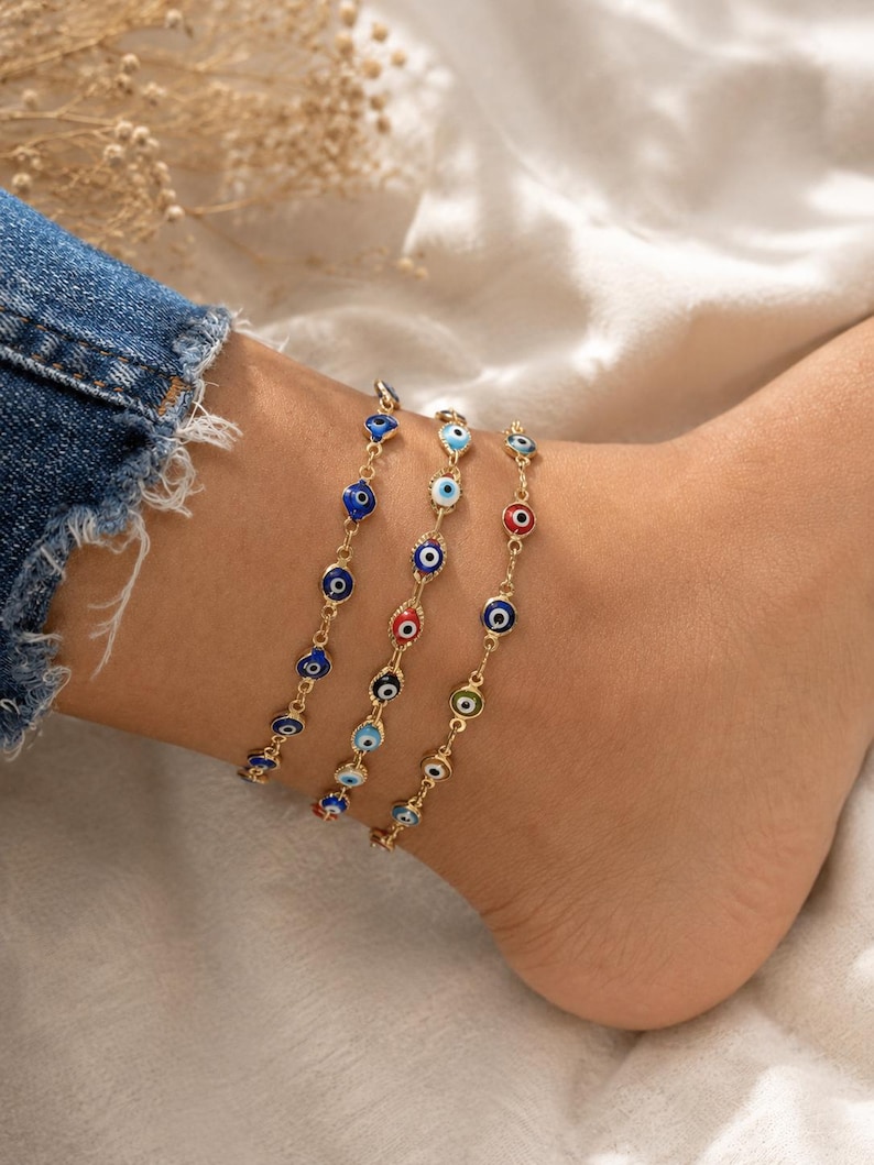 May include: Three gold-tone chain anklets featuring small, circular evil eye charms. The charms are decorated with enamel in various colors, including dark blue, light blue, red, white, and green, arranged in concentric circles on each link.