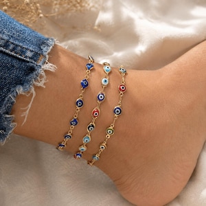May include: Three gold-tone chain anklets featuring small, circular evil eye charms. The charms are decorated with enamel in various colors, including dark blue, light blue, red, white, and green, arranged in concentric circles on each link.
