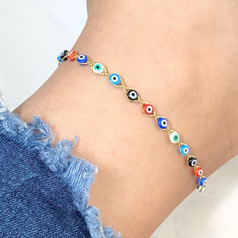 May include: A gold-toned chain anklet featuring a series of small, circular evil eye charms in various colors including red, blue, green, and black. The delicate metal links connect each individual eye charm in a continuous, repeating pattern.