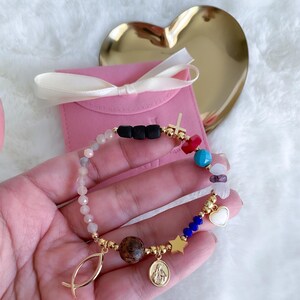 May include: A gold bracelet with a variety of charms, including a cross, a heart, a star, a fish, and a small image of the Virgin Mary. The bracelet is made with small beads of different colors, including pink, blue, red, and white.