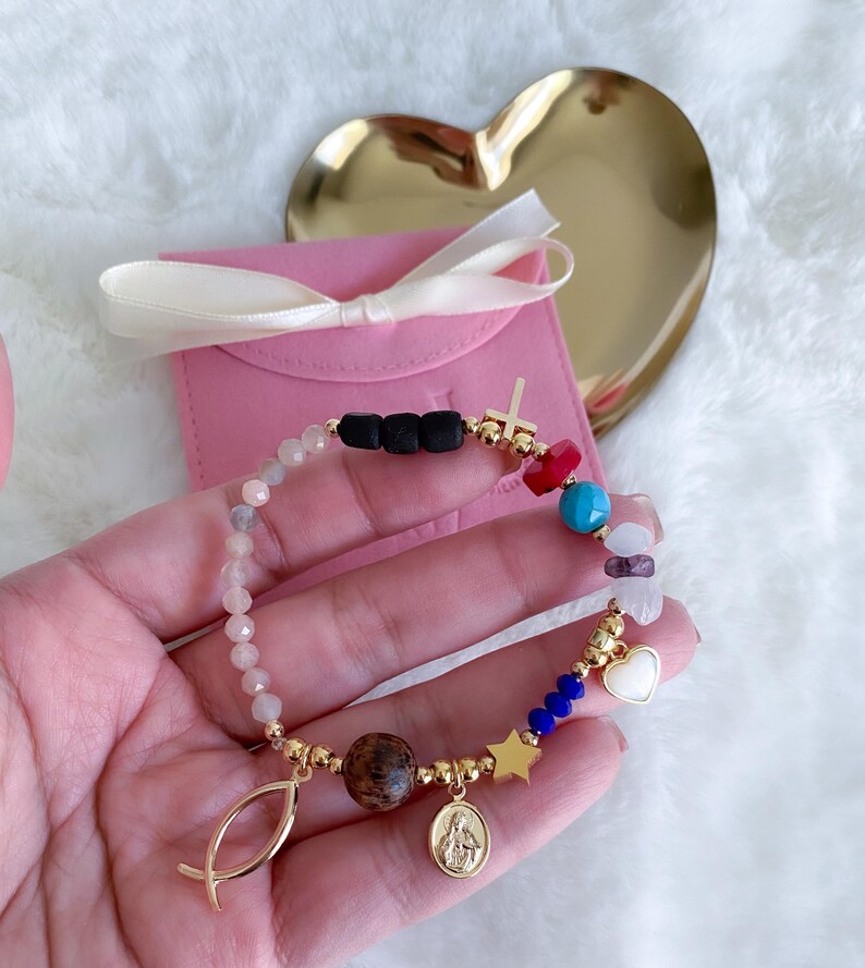 May include: A gold bracelet with a variety of charms, including a cross, a heart, a star, a fish, and a small image of the Virgin Mary. The bracelet is made with small beads of different colors, including pink, blue, red, and white.
