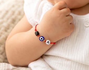 Puede incluir: Una pulsera de hilo rojo con una cuenta de ojo turco azul y blanca, una cuenta negra pequeña y una cuenta redonda blanca con la letra A negra impresa, acentuada por diminutos espaciadores de color dorado en un cordón rojo fino.