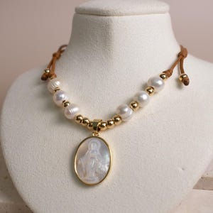 K&ouml;nnte beinhalten: Eine Halskette mit einem zentralen ovalen Anh&auml;nger aus wei&szlig;em Perlmutt, der eine religi&ouml;se Figur darstellt und goldfarben eingefasst ist. Die Kette besteht aus abwechselnden wei&szlig;en S&uuml;&szlig;wasserperlen und kleinen goldenen Perlen an einer braunen Kordel.