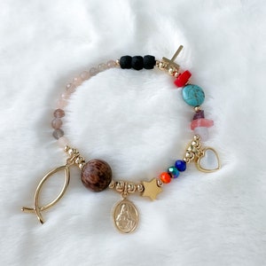 May include: A gold bracelet with a variety of charms, including a cross, a heart, a star, a fish, and a wooden bead. The bracelet also features turquoise, pink, and orange beads.