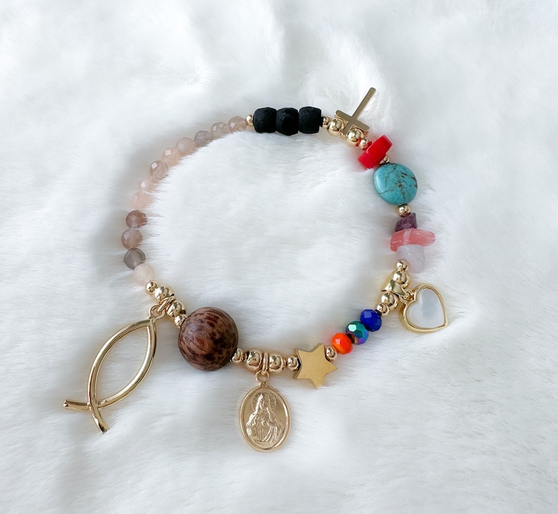 May include: A gold bracelet with a variety of charms, including a cross, a heart, a star, a fish, and a wooden bead. The bracelet also features turquoise, pink, and orange beads.