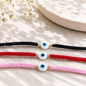 K&ouml;nnte beinhalten: Drei geflochtene Kordelarmb&auml;nder in Schwarz, Rot und Hellrosa. Jedes Armband hat in der Mitte eine wei&szlig;e Nazar-Auge-Perle mit blauer Iris und schwarzer Pupille, die von zwei kleinen goldfarbenen Metallperlen auf jeder Seite flankiert wird.