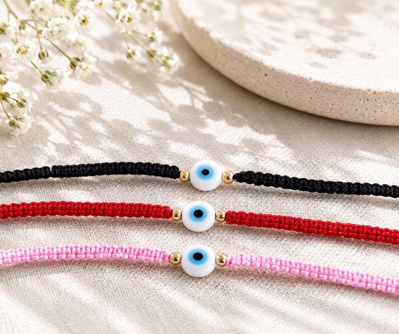 K&ouml;nnte beinhalten: Drei geflochtene Kordelarmb&auml;nder in Schwarz, Rot und Hellrosa. Jedes Armband hat in der Mitte eine wei&szlig;e Nazar-Auge-Perle mit blauer Iris und schwarzer Pupille, die von zwei kleinen goldfarbenen Metallperlen auf jeder Seite flankiert wird.